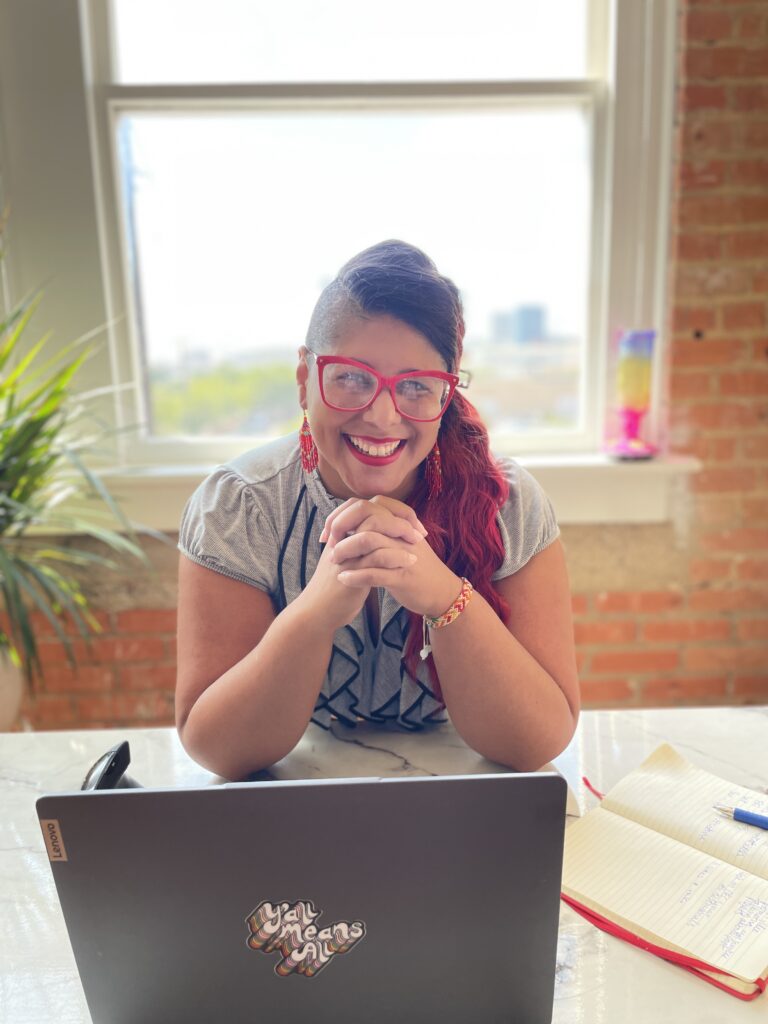 Photo of Jesenia at a marble desk sitting in front of a window, set in a brick wall. At the desk Jesenia is clasping her hands together under her chin in front of a laptop and mouse. The laptop has a sticker that reads "Y'all means All".  There is a palm plant next to the desk and a city skyline blurred in the background through the window. It is early afternoon with sun coming through the window.  Jesenia is an indigenous woman with a red faux hawk in a twist, wearing pink glasses, gold red native earrings and red lipstick with a grey ruffle neck blouse. 