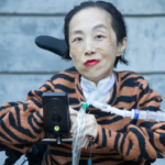 Photo of Alice Wong, an Asian American disabled woman in a power chair. She is wearing a black blouse with a floral print, a bold red lip color and a trach at her neck. She is giving a cheeky expression with her eyebrow partially raised. In the background is a gray cement wall. Photo credit: Eddie Hernandez Photography.