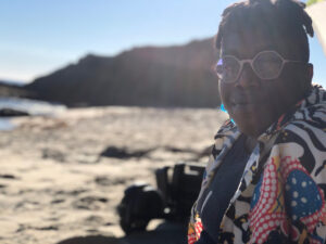 Bri smiles slightly at the camera while wearing a multi-patterned shirt, hexagonal purple glasses and blue glass earrings. Behind zir is sand, ocean waves and a jagged cliff.
