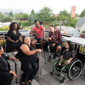 Overhead shot of six disabled people of color at a rooftop deck party. An Indigenous Two-Spirit person with a prosthetic leg smiles directly at the camera and gives a thumbs up while everyone else is engaged in conversation.