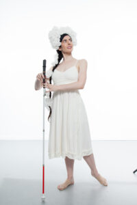 Krishna, a thin, pale skinned woman with long dark hair pulled into a braid, poses on a studio stage. She is wearing a simple white dress with her shoulders exposed and a fluffy white headdress perched on her forehead and wrapping down her braid. She looks over her left shoulder as she lightly holds a straight cane between her hands and points her left foot behind her. Photo Credit: Krishna Washburn & Micaela Mamede